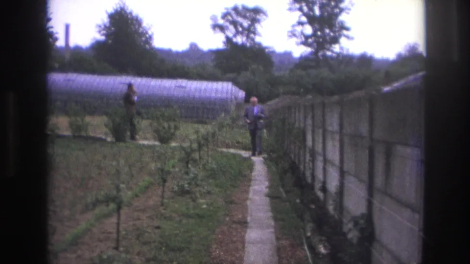 Paris, France, 1967 frame with a suited man on a garden path, another man near the greenhouse, and crop rows nearby.