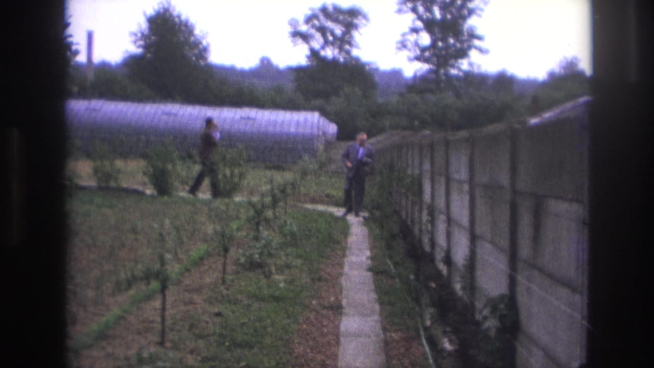 1967 Paris home-movie frame showing two adult men in a garden beside a concrete path, greenhouse, planted rows, and tall wall.