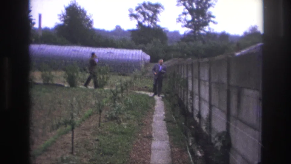 1967 Paris home-movie frame showing two adult men in a garden beside a concrete path, greenhouse, planted rows, and tall wall.