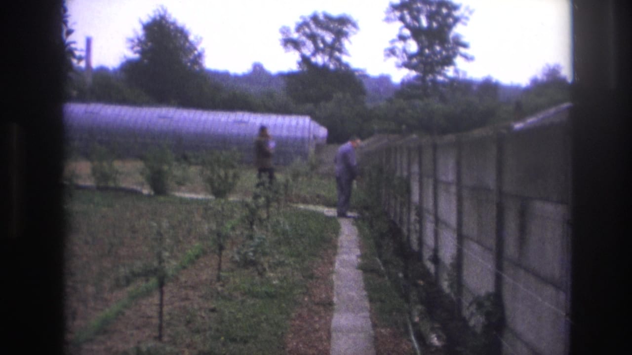 Two men near a garden path and wall with a greenhouse in the background.
