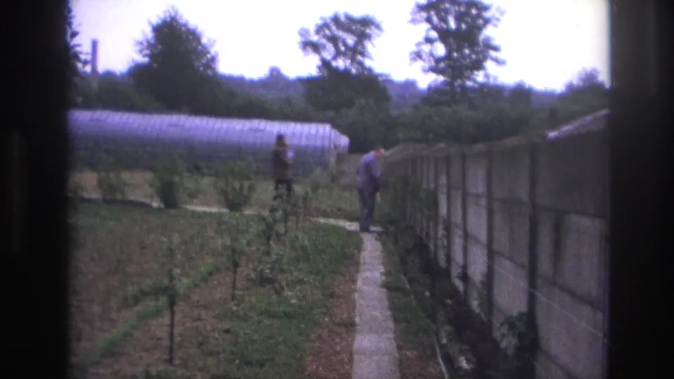 1967 Paris home-movie frame with two adult men moving near a narrow path, concrete wall, planted garden, and greenhouse.