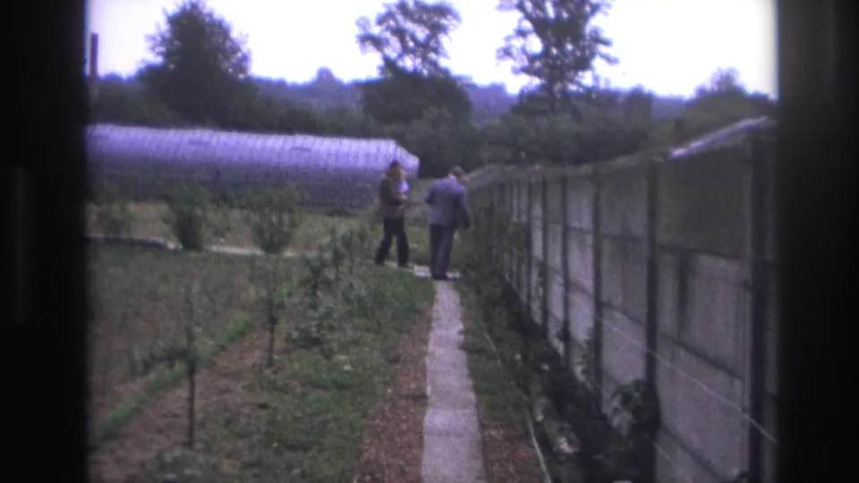 1967 Paris home-movie frame showing two adult men near each other on a garden path, with greenhouse, wall, and planted rows visible.