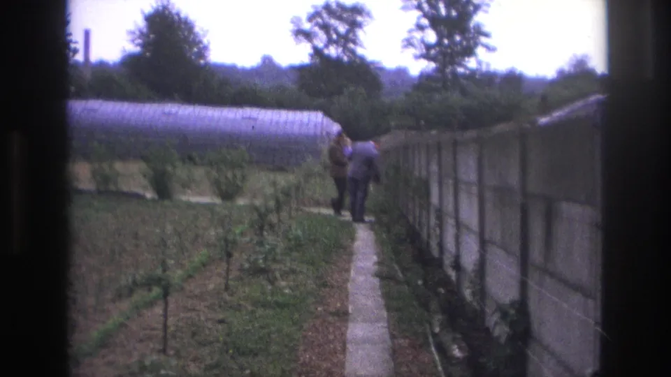 1967 Paris home-movie frame of two adult men close together on a garden path, with a greenhouse and concrete wall in view.
