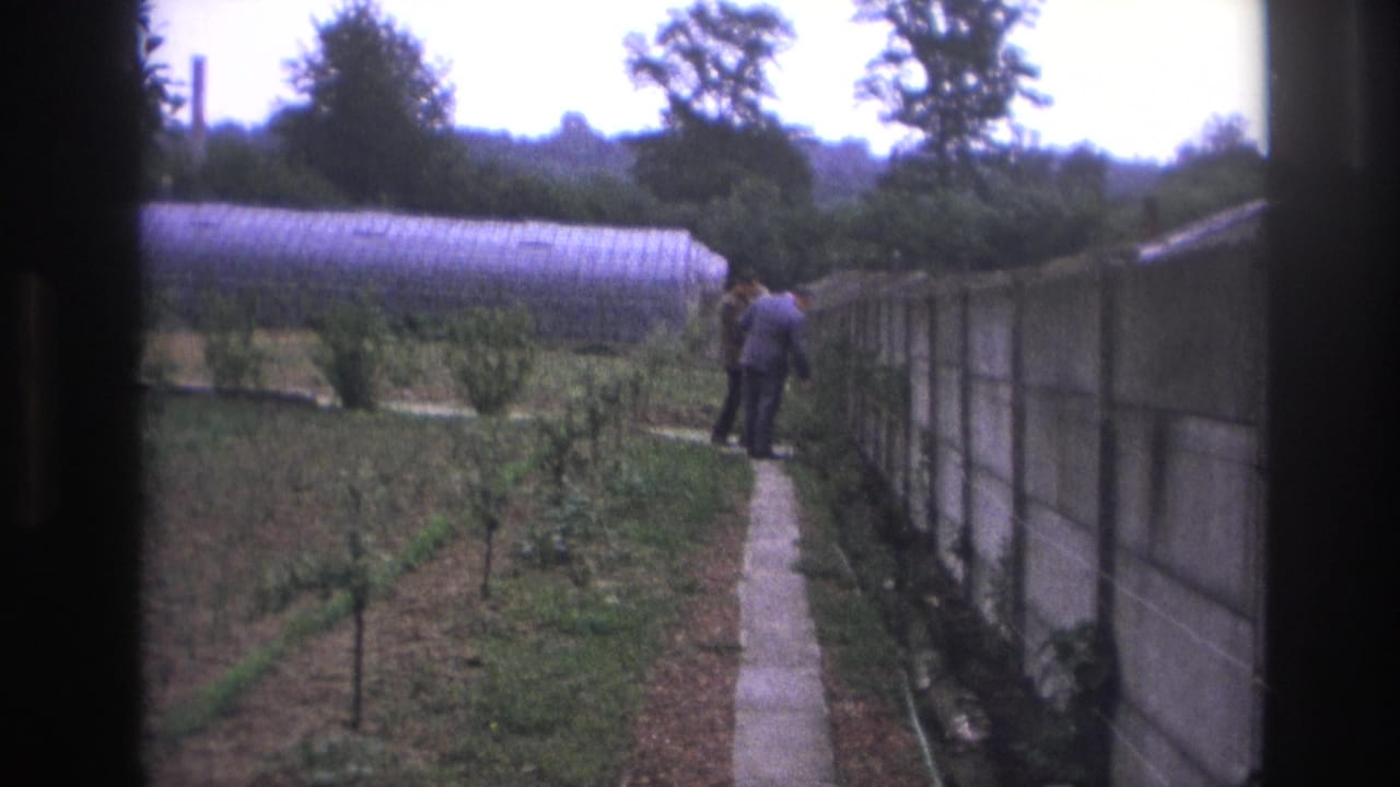 Two suited men stand on a narrow path beside plantings and greenhouse structures.