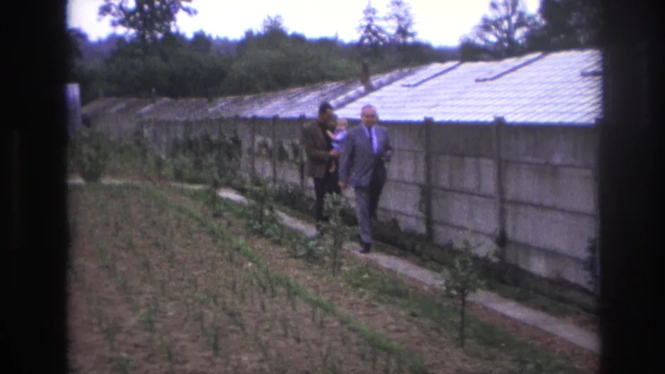 A man in a light suit and another in a dark jacket walk toward the camera along a narrow greenhouse-side path in Paris, France, 1967.