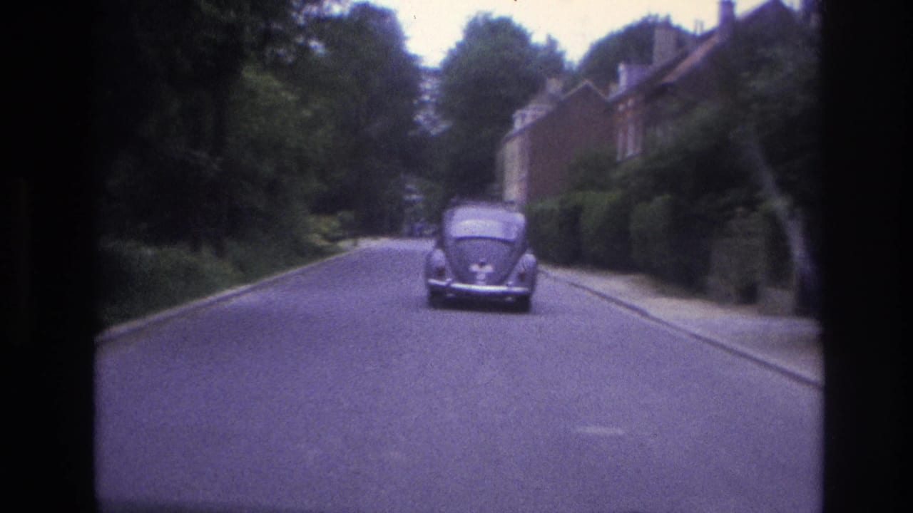 1967 Paris 8mm frame showing a Volkswagen Beetle driving ahead on a tree-lined residential street with brick houses and hedges.