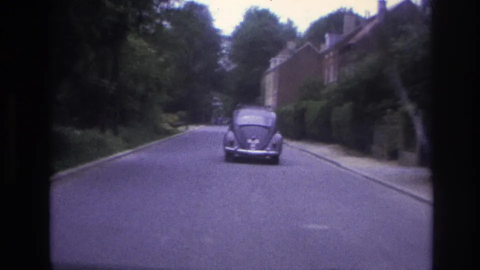 1967 Paris 8mm frame showing a Volkswagen Beetle driving ahead on a tree-lined residential street with brick houses and hedges.