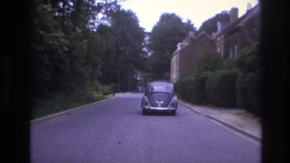 Rear view of a Volkswagen Beetle on a quiet Paris residential street, with trees, hedges, and brick buildings visible through 8mm grain.