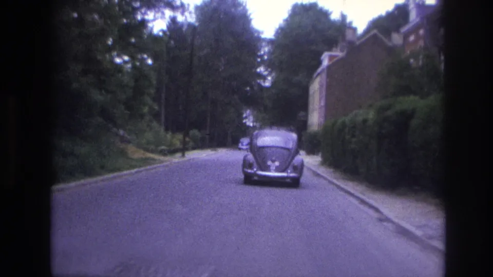 1967 Paris street view with a Volkswagen Beetle centered ahead, brick homes and trimmed hedges on the right, and trees along the roadway.