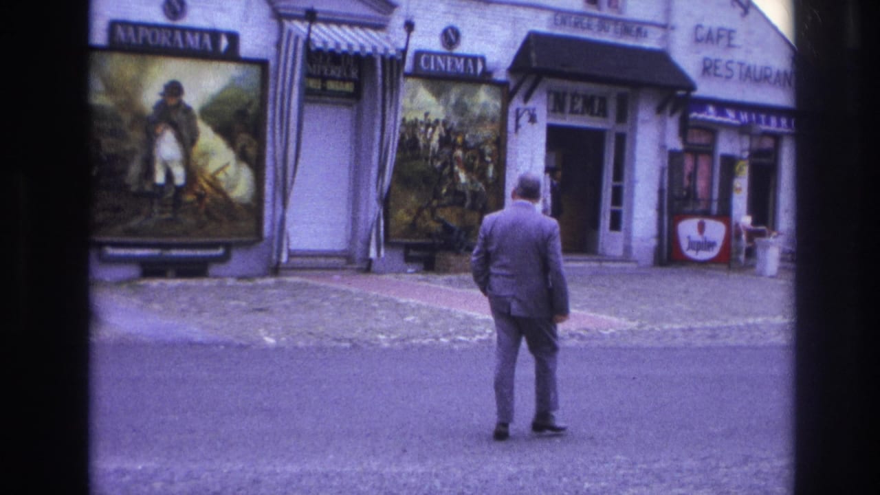 In 1967 Paris, a suited man pauses or steps from the street toward a cinema entrance beside cafe-restaurant signage and large painted movie posters.