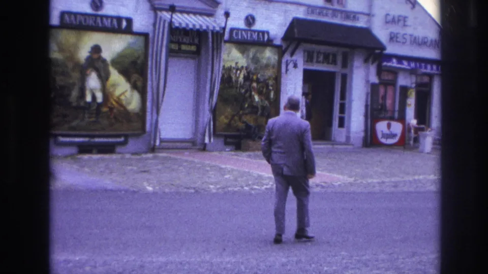 In 1967 Paris, a suited man pauses or steps from the street toward a cinema entrance beside cafe-restaurant signage and large painted movie posters.