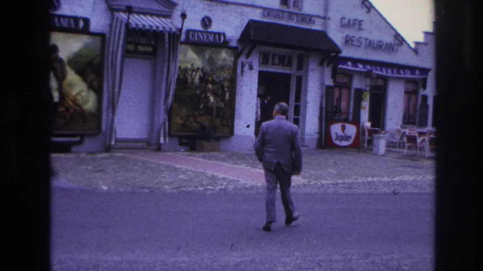 A suited pedestrian crosses a Paris street toward a cinema and cafe frontage, with French signage, poster displays, and outdoor seating visible.