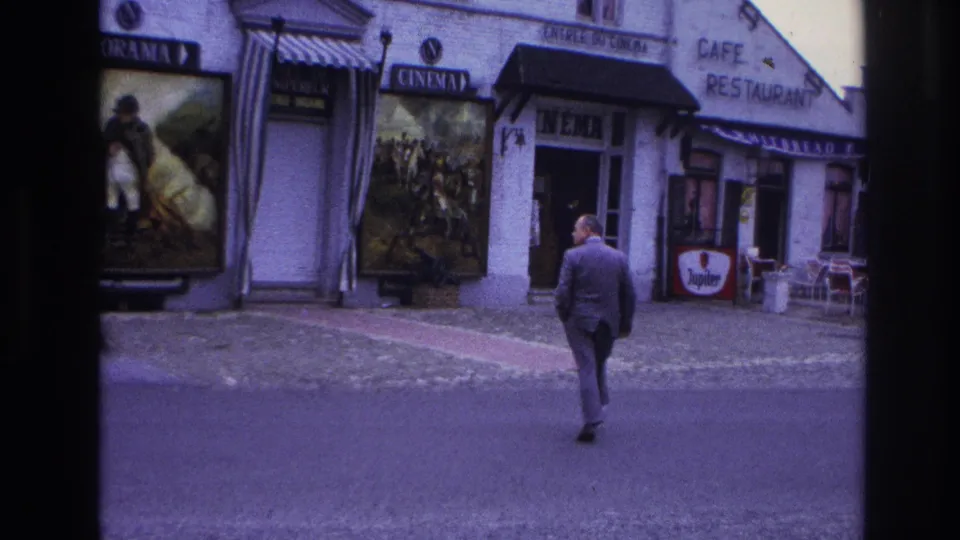 The man in a gray suit moves closer to the cinema entrance as the cafe frontage, poster boards, striped awning, and street paving remain in view.