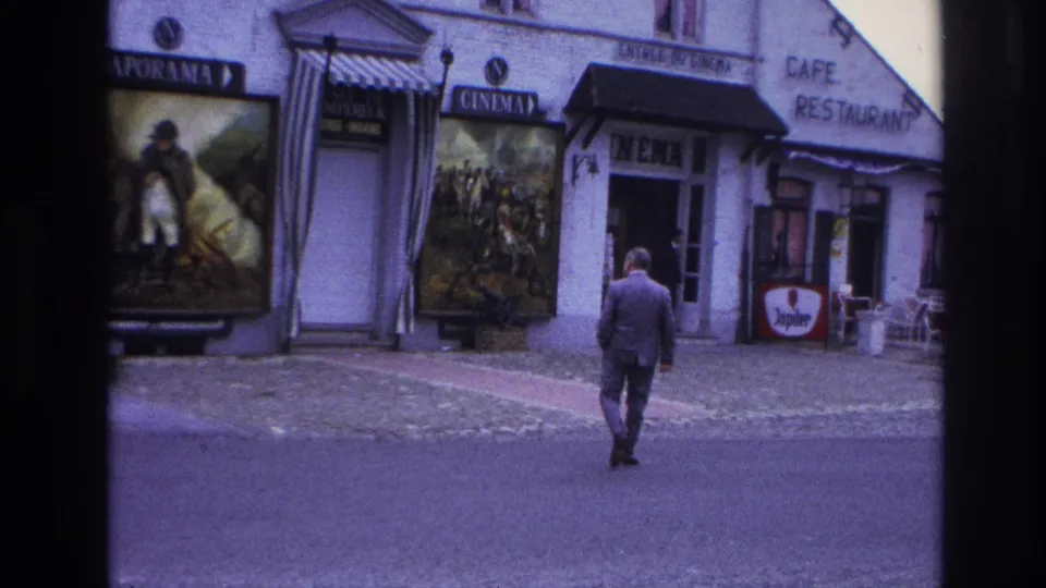 At the end of the shot, the suited man nears the sidewalk in front of the Paris cinema and cafe, with poster displays and outdoor seating still visible.