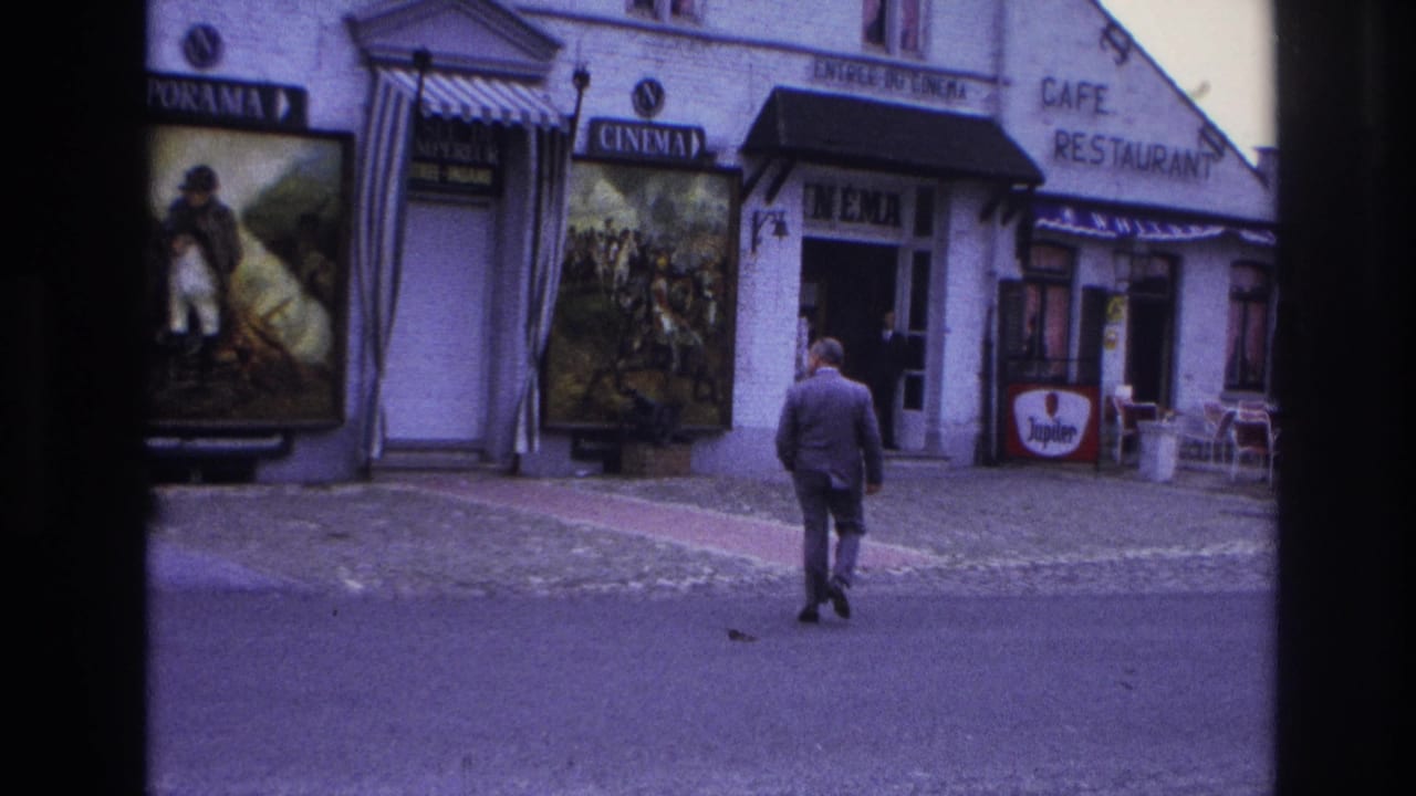 1967 Paris home-movie frame showing a suited man crossing a quiet street toward a cinema entrance beside a cafe-restaurant.