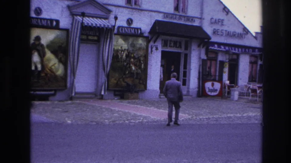 1967 Paris frame with a suited man paused near the curb, facing a cinema facade with posters and neighboring cafe seating.