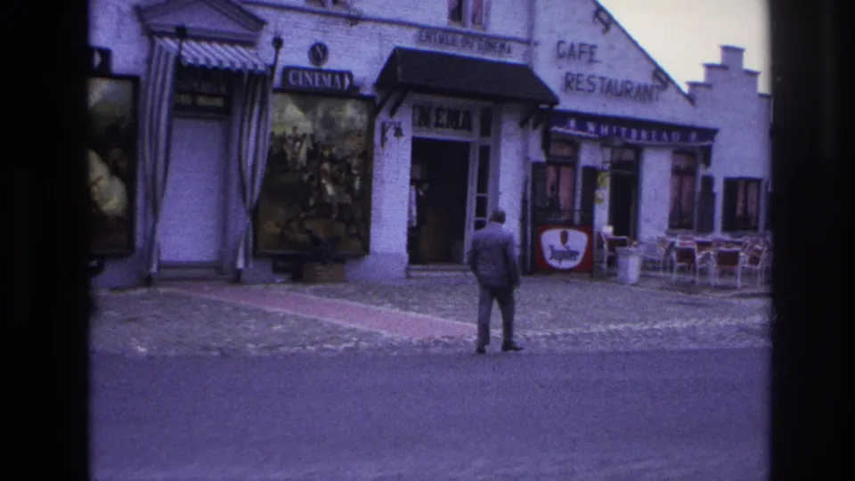 1967 Paris street view showing the suited man centered before a cinema, with cafe-restaurant signage and chairs to the right.