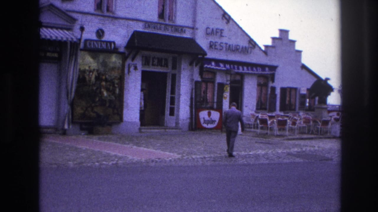 Suited man walking past the cinema entrance and cafe seating beside a beer sign.