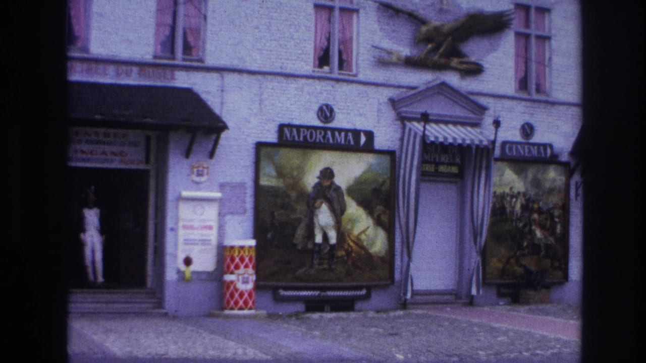 Building entrance with a person in white standing near Naporama posters, cinema signs, awnings, and barrels.