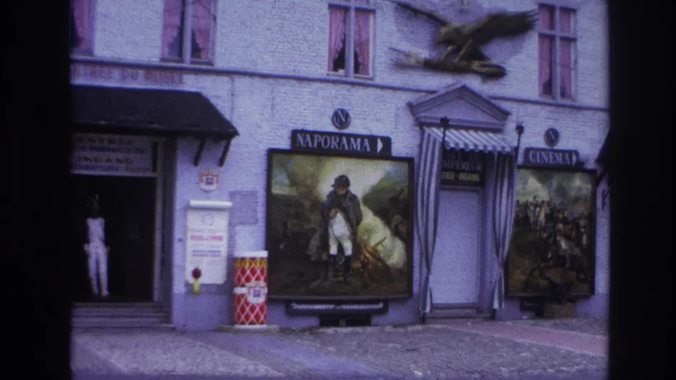 1967 Paris 8mm frame of the Naporama building showing a doorway figure in white, large posters, cinema signage, striped awning, and red barrels.