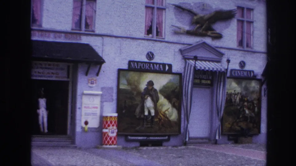 1967 Paris 8mm frame with a doorway figure beside a large Naporama soldier poster, cinema sign, striped awning, eagle sculpture, and facade details.