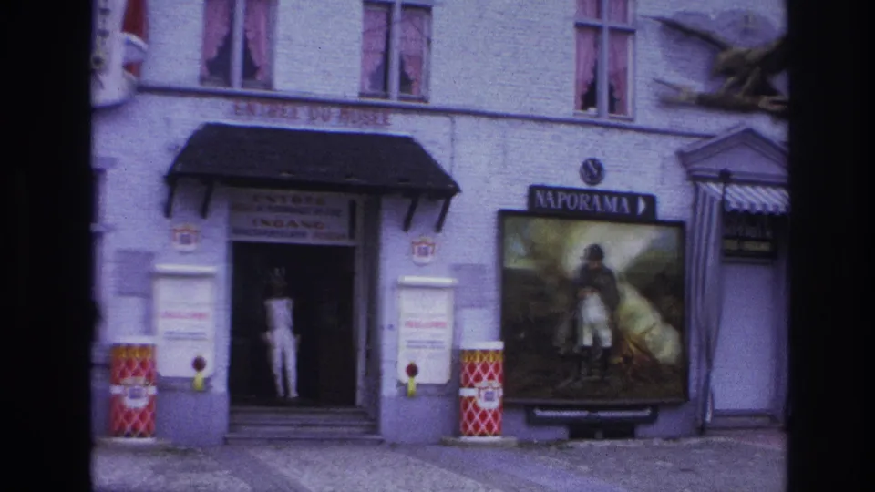 1967 Paris 8mm frame showing the Naporama facade with a doorway figure, soldier poster, red patterned barrel, striped awning, and eagle sculpture overhead.