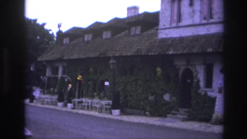 1967 Paris 8mm frame with a darkened pan across an ivy-covered facade, arched entrance, outdoor seating, and people near the walkway.