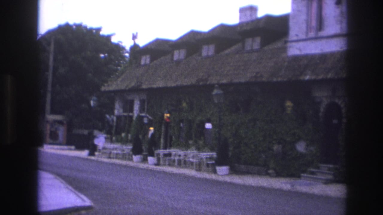 1967 Paris home-movie frame showing an ivy-covered street building, cafe tables, sidewalk planters, and a curved roadway.