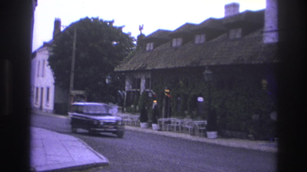 Dark car passing an ivy-covered building with outdoor seating on the sidewalk.
