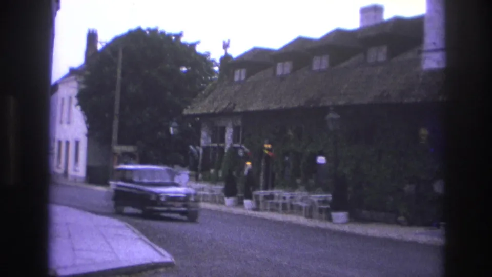 1967 Paris street view with a dark mid-century car moving past an ivy-covered building and sidewalk cafe seating.