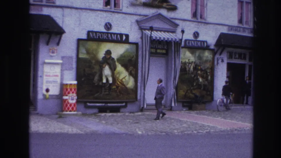 1967 Paris home-movie frame of a cinema exterior with large posters, a man walking, a bicycle, signage, and cobblestone paving.