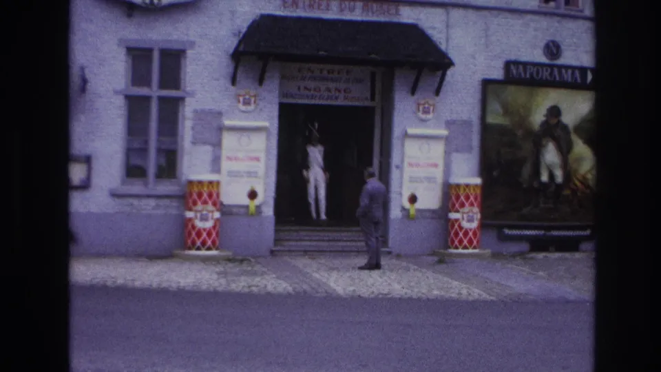 A man stands with his back to the camera outside a Paris museum entrance, facing the doorway and adjacent Naporama poster.