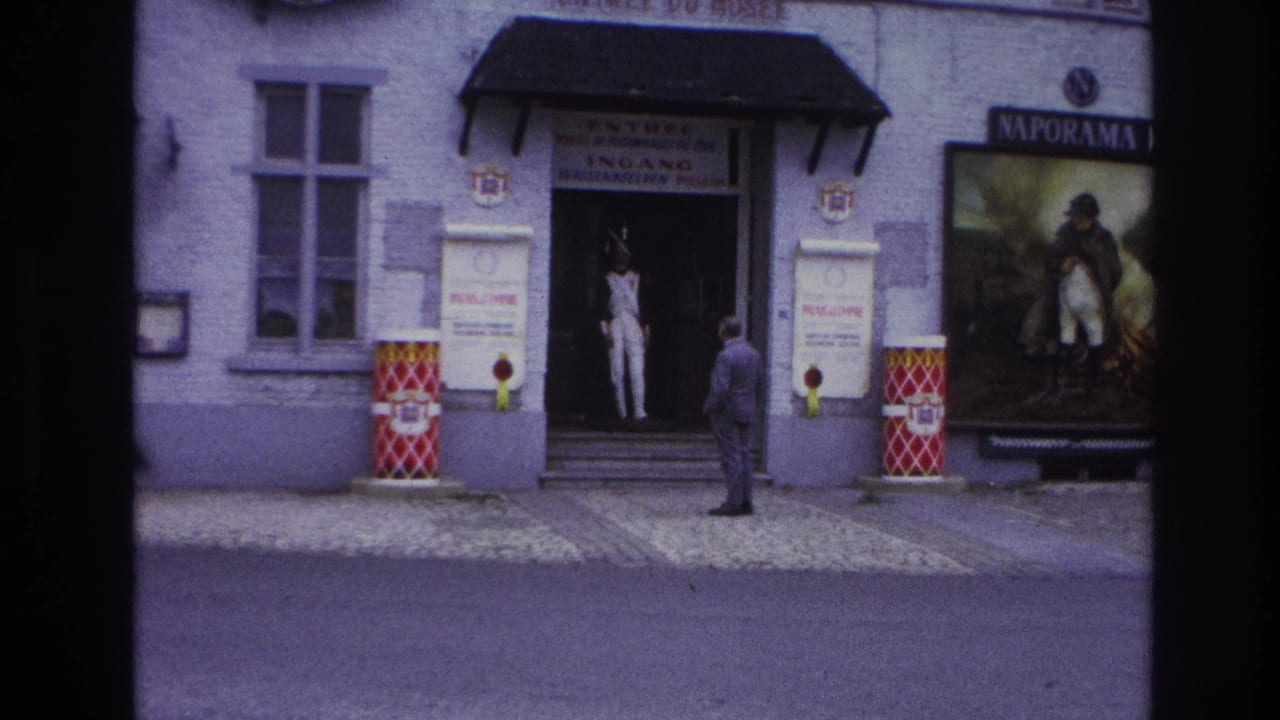 1967 Paris home-movie frame showing a public entrance, a suited man facing the doorway, a white-uniformed figure inside, and poster signage.