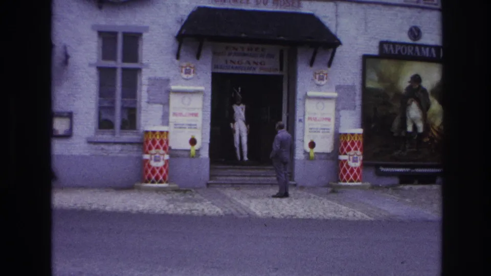 1967 Paris home-movie frame showing a public entrance, a suited man facing the doorway, a white-uniformed figure inside, and poster signage.