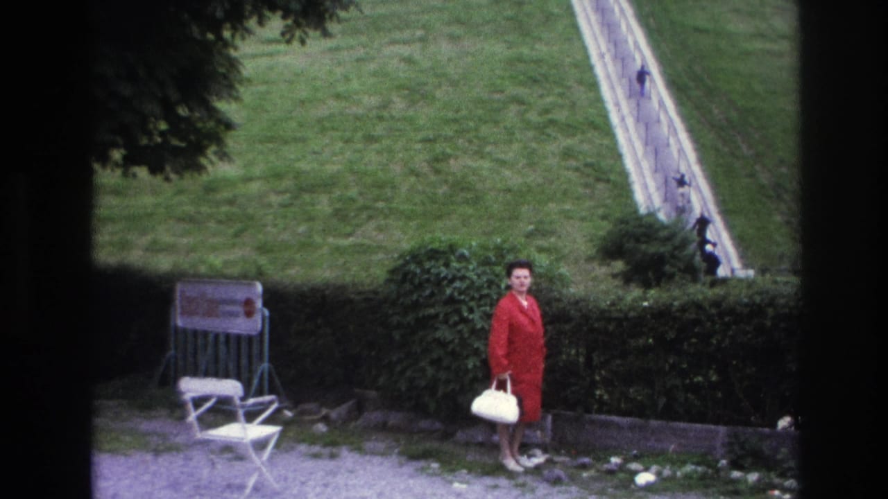 Woman in a red coat stands near a hedge with a white handbag, lawn, folding chair, and sloped pathway behind her.