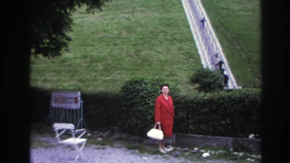 1967 Paris home-movie frame showing a posed woman in a red coat looking toward the camera beside a hedge and sloping garden path.