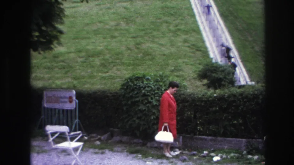 1967 Paris home-movie frame of the woman in a red coat turning downward near the hedge, with lawn and a long sloped path in the background.