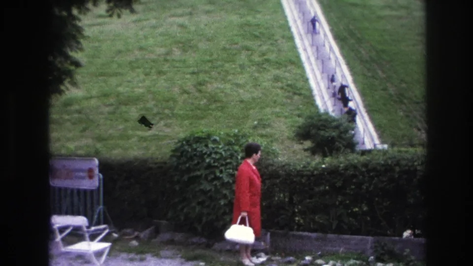 1967 Paris 8mm frame showing a woman with a white handbag watching distant pedestrians climb a steep white stairway across a grassy hill.