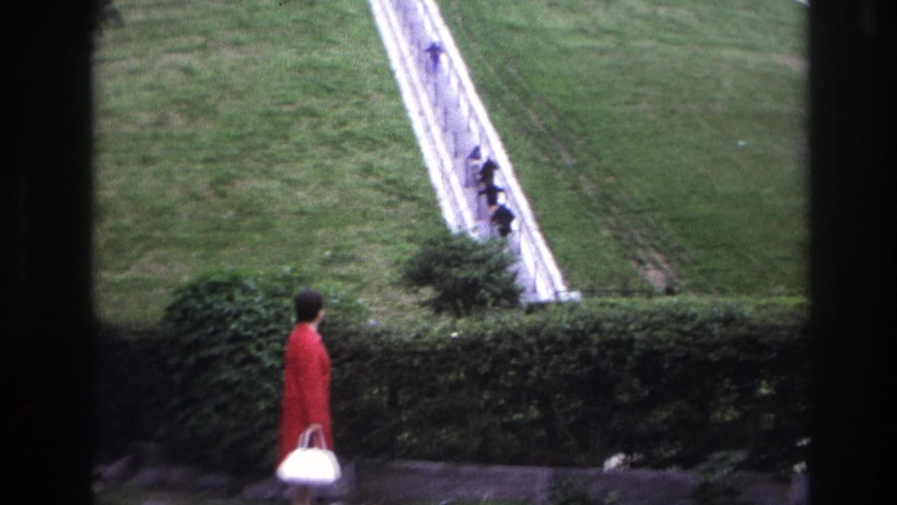 1967 Paris home-movie frame with a woman in red seen near a hedge, looking toward small figures ascending a steep stairway on grass.