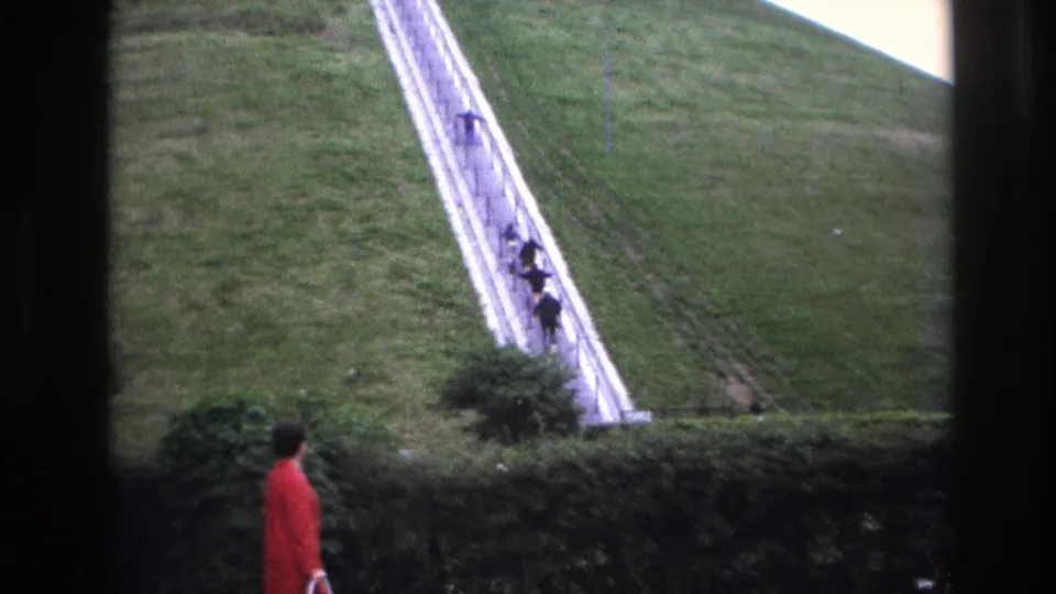 1967 Paris 8mm frame showing the woman partly lower in view, with the long pale stairway and distant visitors rising behind the hedge.