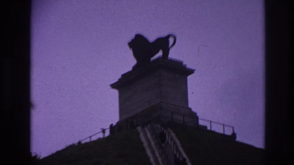 1967 Paris home-movie frame showing a silhouetted lion statue atop a stone pedestal, with railing and monument edges below.