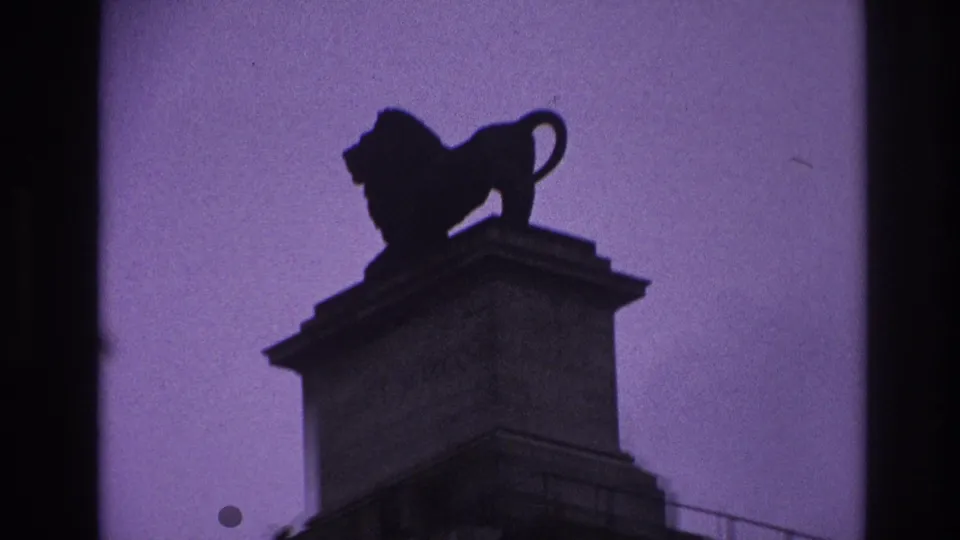 1967 Paris home-movie still showing a closer, slightly soft view of a lion statue on a high stone pedestal.