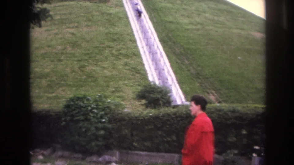 1967 Paris home-movie frame showing the woman in red walking past a hedge below the long white hillside staircase.