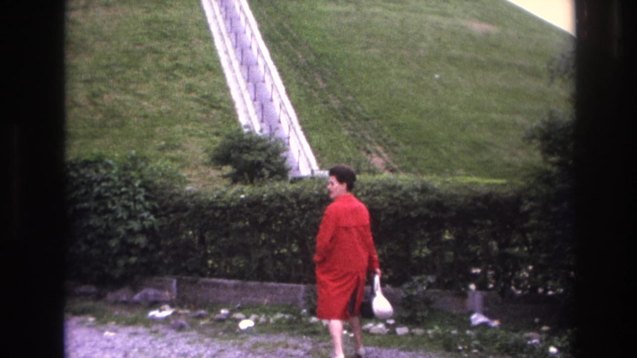 In 1967 Paris home-movie footage, a woman in a red coat walks along a gravel path beside a hedge and grassy slope.