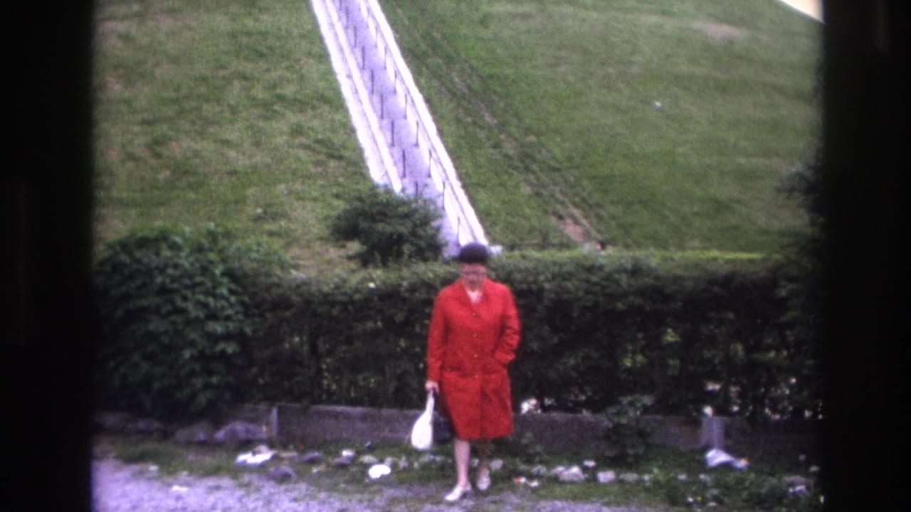 In 1967 Paris home-movie footage, a woman in a red coat stands by a hedge near a grassy slope and pale railing.
