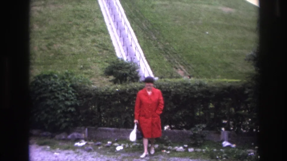 In 1967 Paris home-movie footage, a woman in a red coat stands by a hedge near a grassy slope and pale railing.
