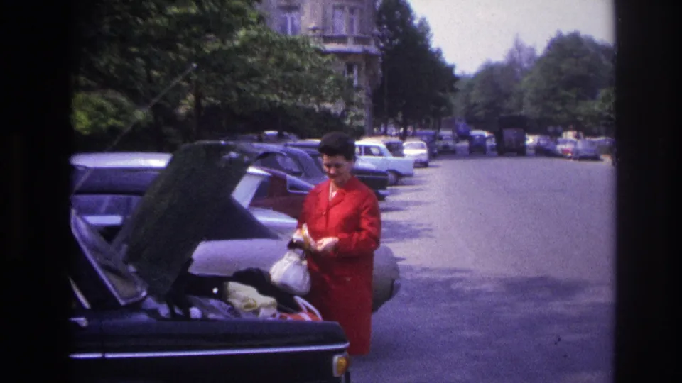 A 1967 Paris home-movie frame shows the woman beside a dark car with its trunk open, with parked cars stretching along the street.