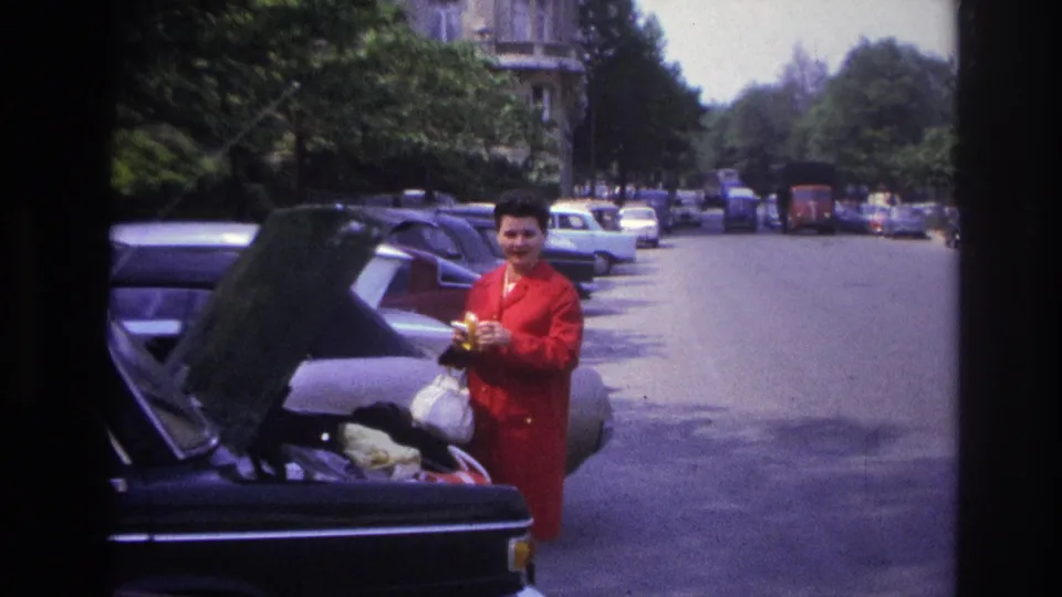 In 1967 Paris home-movie footage, the woman smiles beside the open trunk while holding a banana and white bag on a parked-car-lined street.