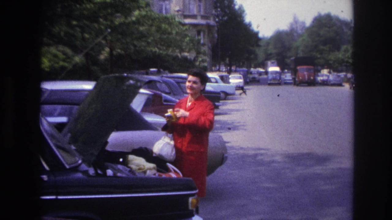 In 1967 Paris home-movie footage, a woman in a bright red coat stands by an open car boot with parked cars and traffic behind her.