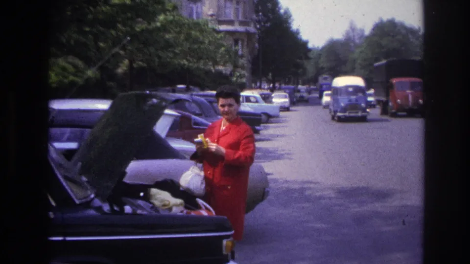 The woman stands near the open boot and faces the camera, with a blue truck, a red vehicle, and rows of cars visible down the Paris street.
